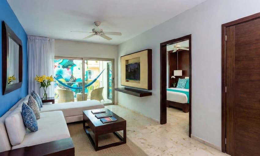 Sala de suite con sofá blanco, mesa de madera, televisión y balcón con hamaca azul.