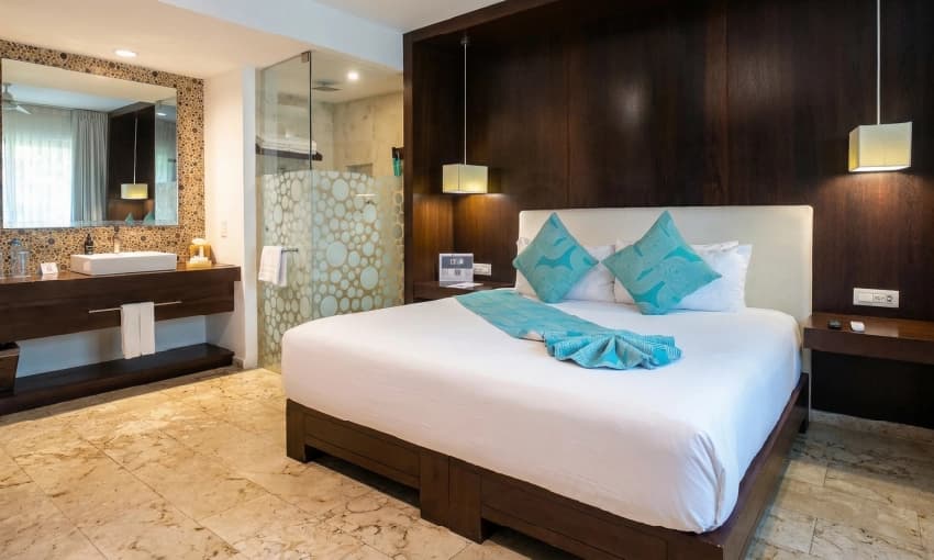 Habitación con cama king, cabecera de madera oscura, cojines turquesas y área de lavabo.