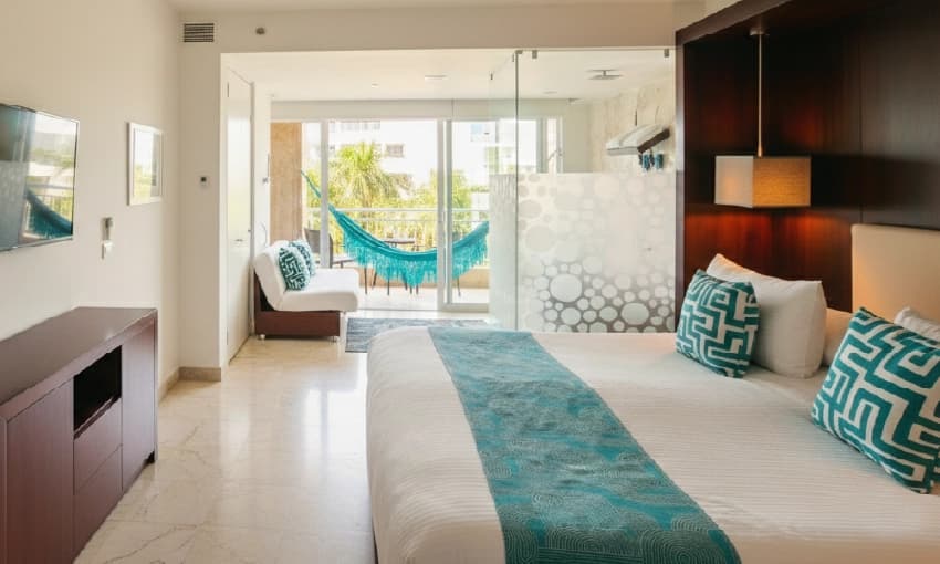 Habitación tipo loft con cama blanca, detalles turquesas y terraza con vista a palmeras.