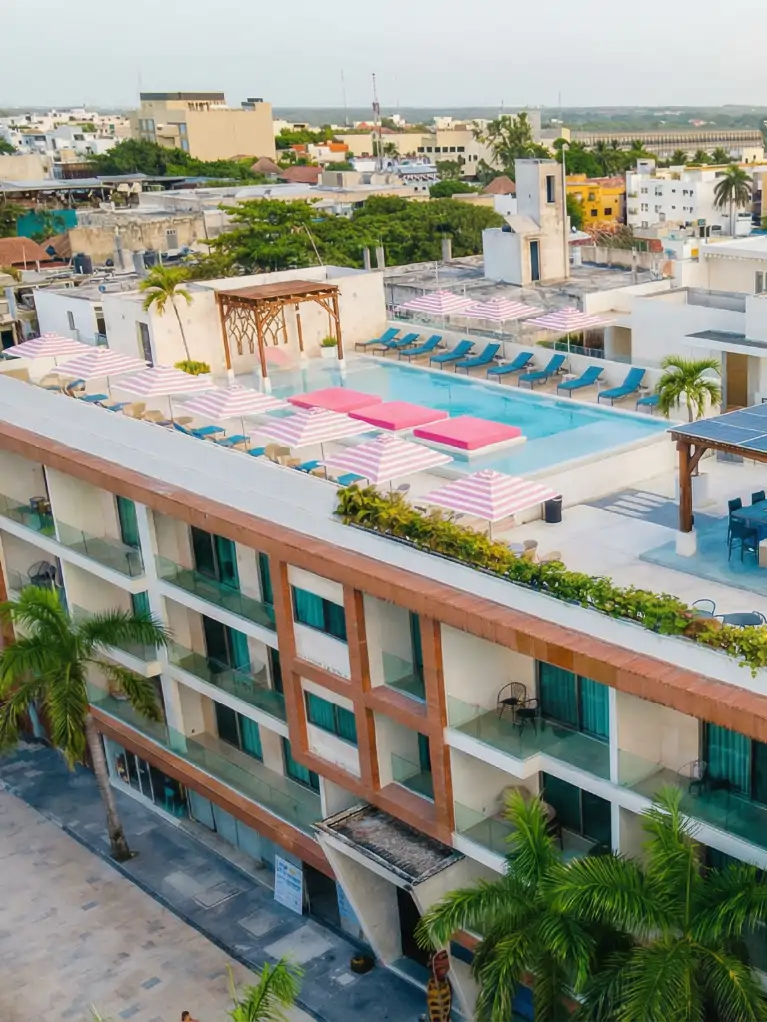 Terraza de hotel con alberca, camastros azules y sombrillas de rayas rosas frente a la ciudad.