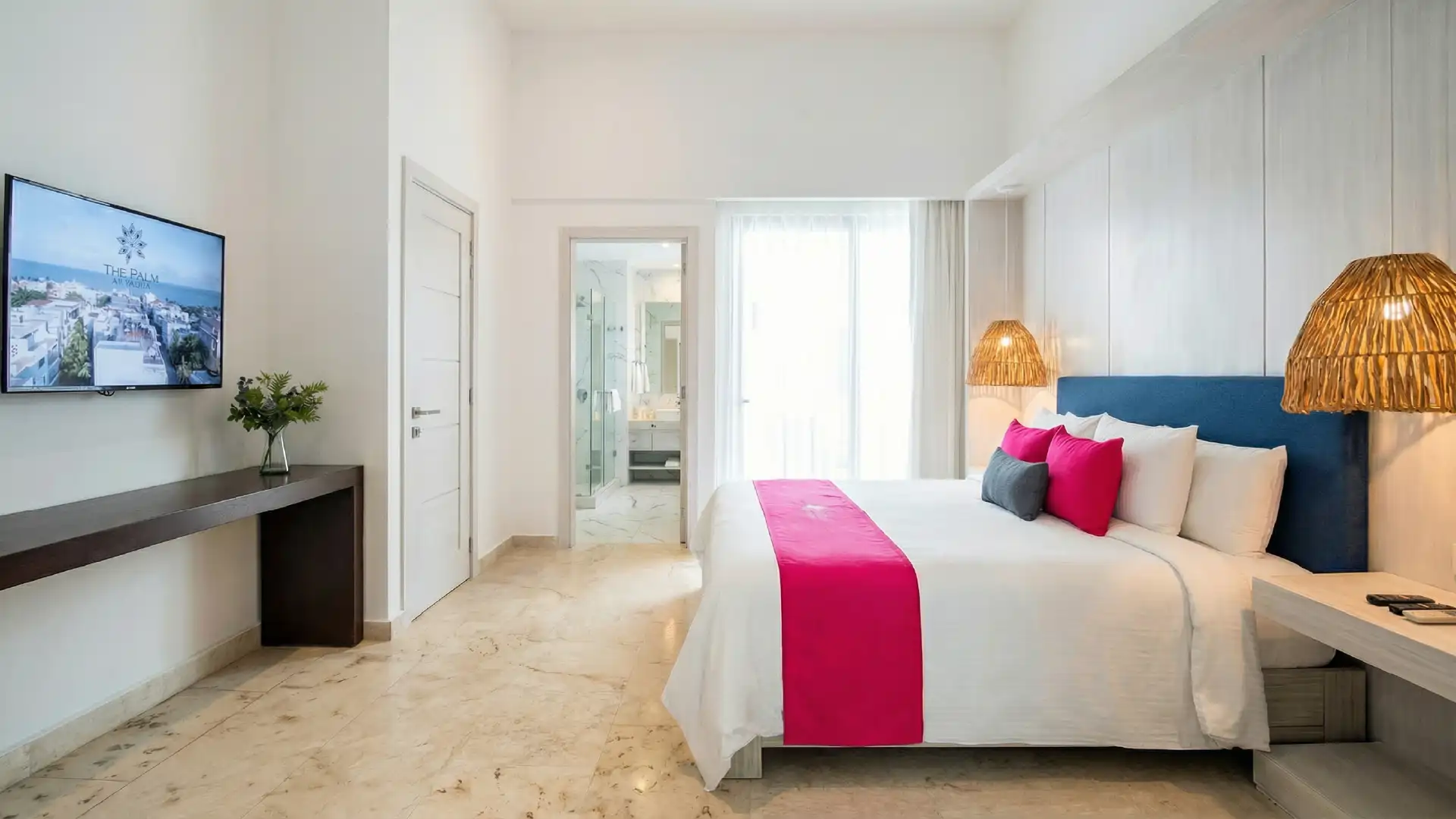 Habitación de hotel moderna con cama blanca, cojines rosas, lámparas de mimbre y televisión.