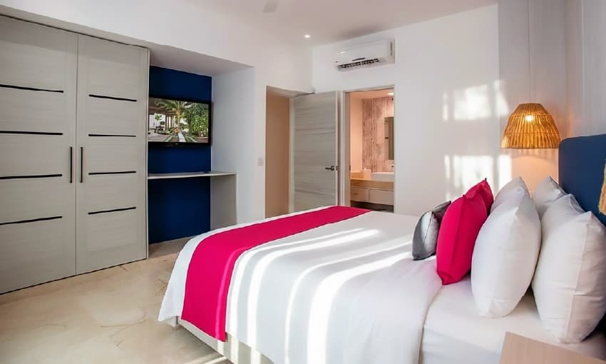 Suite moderna con cama amplia, cojines rosas fucsia, clóset minimalista y aire acondicionado.