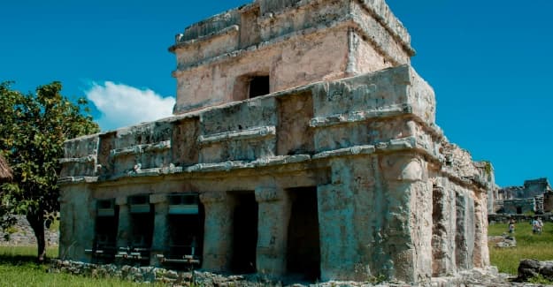 Ruinas mayas de piedra en una zona arqueológica bajo un cielo azul despejado y pasto verde.